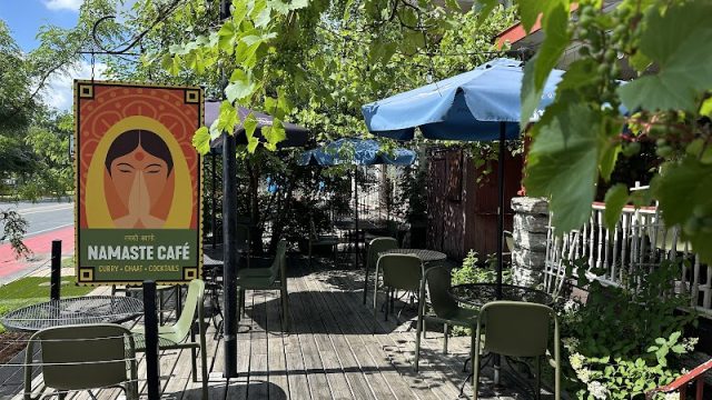 Outdoor patio seating at Namaste Café in Uptown Minneapolis, with colorful café signage, umbrellas, tables and chairs surrounded by greenery.