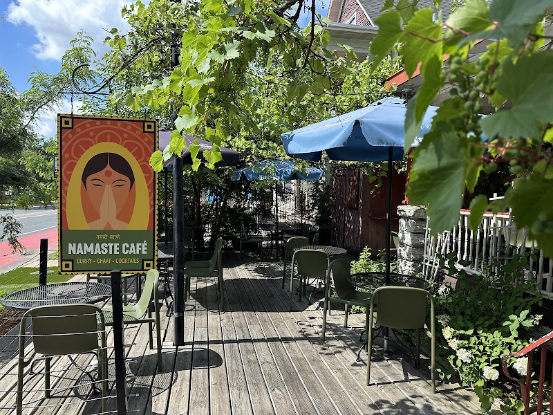 Outdoor patio seating at Namaste Café in Uptown Minneapolis, with colorful café signage, umbrellas, tables and chairs surrounded by greenery.